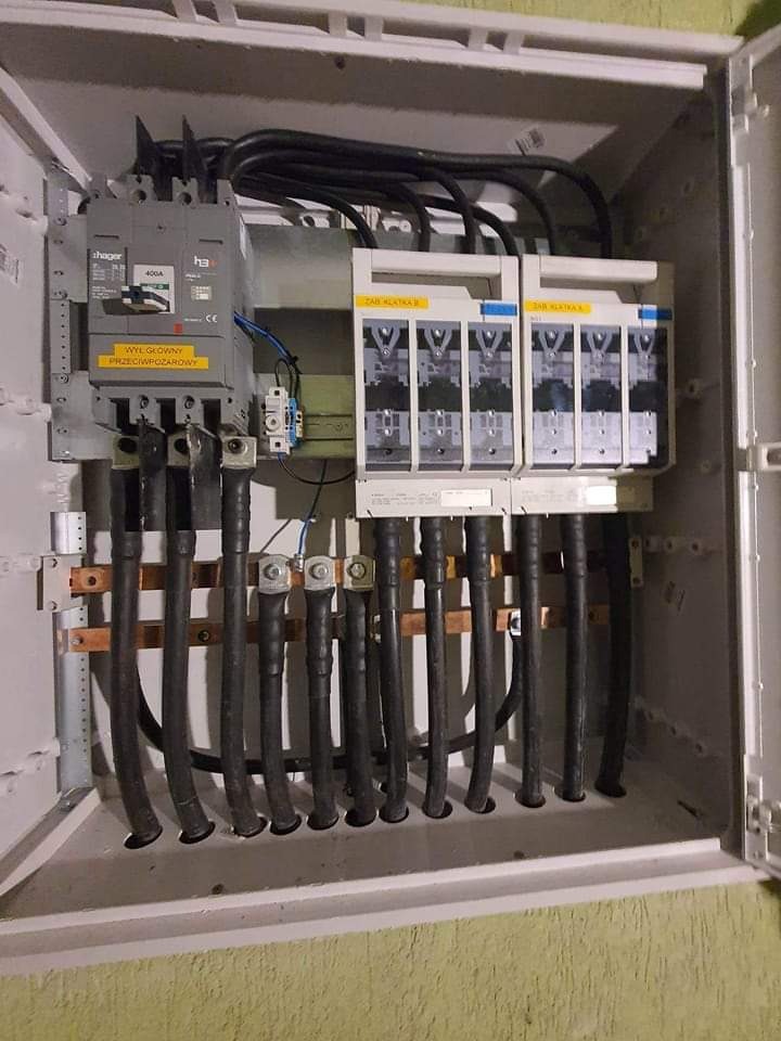 Otwarta skrzynka rozdzielcza z zamontowanymi bezpiecznikami, wyłącznikiem głównym przeciwpożarowym i grubymi kablami elektrycznymi.