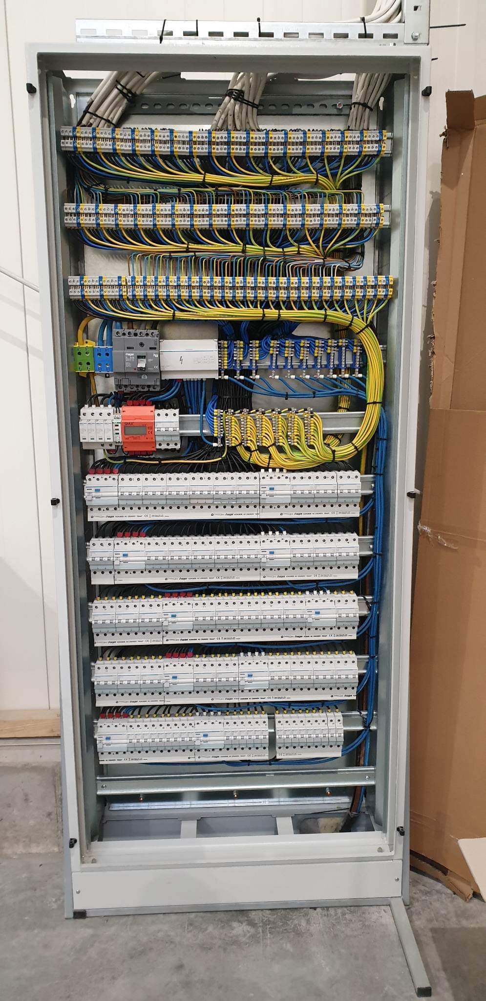 Rozdzielnica elektryczna z licznymi zabezpieczeniami i okablowaniem w kolorach niebieskim i żółtym, widoczne bezpieczniki i listwy zaciskowe.