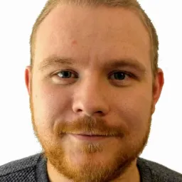 Portret mężczyzny z rudą brodą i krótkimi blond włosami, ubrany w szary sweter o wyraźnej fakturze.