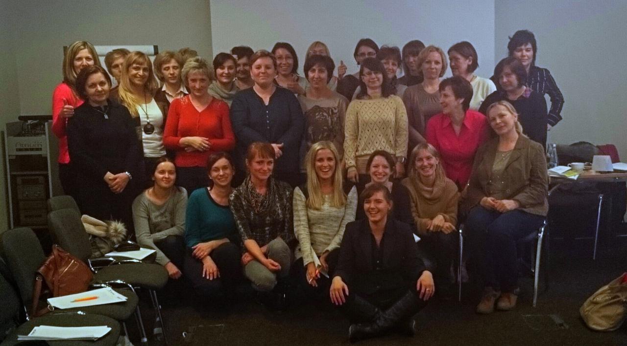 Grupa uśmiechniętych kobiet w sali konferencyjnej, niektóre siedzą na krzesłach, inne na podłodze, z notatkami i długopisami na krzesłach.