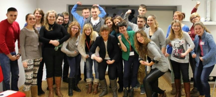 Grupa uśmiechniętych osób, w różnym wieku, stojących w pomieszczeniu i pozujących z podniesionymi pięściami, symbolizującymi siłę i sukces, widoczna kobieta w zielonej marynarce.