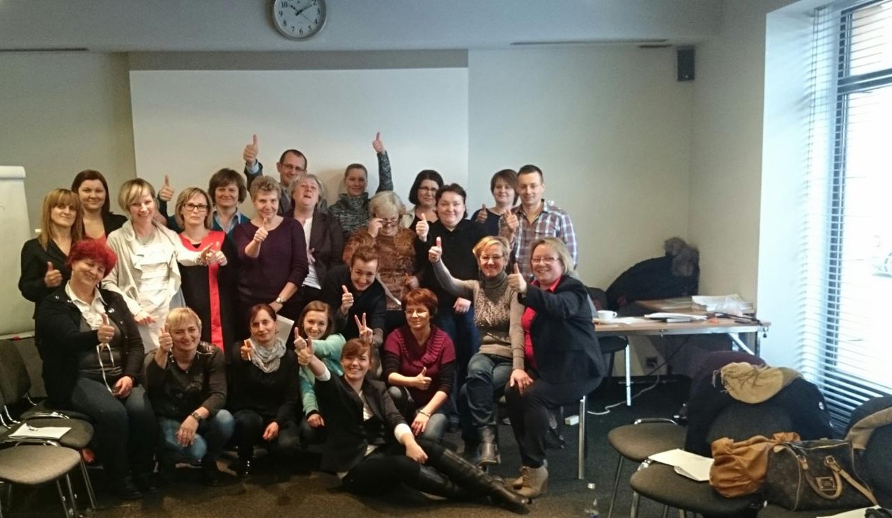 Grupa uśmiechniętych osób, uczestników kursu języka angielskiego, pozuje z podniesionymi kciukami w sali z zegarem na ścianie i stolikiem z notatkami w tle.