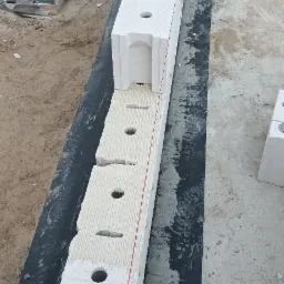 Wznoszenie ściany z betonu komórkowego Ytong: widoczny pierwszy rząd bloczków z otworami, ułożonych na czarnej hydroizolacji, z naniesioną zaprawą, obok worki z zaprawą murarską Silmur M-7,5.