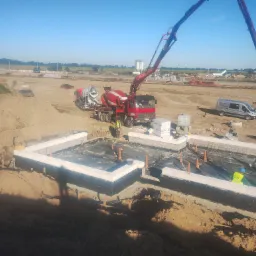 Wykop pod fundamenty z hydroizolacją, w tle betoniarka z pompą podająca beton. Widoczni pracownicy w odblaskowych kamizelkach. Słoneczny dzień na placu budowy.