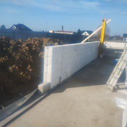 Murtechnik Paweł Żurek - Wznoszenie ściany z białych bloczków betonowych na fundamencie. Obok leży pryzma ziemi i stoi drabina. Widoczne elementy szalunku. Słoneczny dzień.