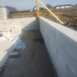 Murtechnik Paweł Żurek - Wznoszenie ścian fundamentowych z bloczków betonu komórkowego. Widoczny fragment ściany, poziomica, palety bloczków i samochód dostawczy na tle krajobrazu.