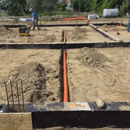 Murtechnik Paweł Żurek - Plac budowy z wykopami pod instalacje, ułożonymi pomarańczowymi rurami kanalizacyjnymi i pracownikiem zagęszczającym grunt. Widoczne zbrojenie.