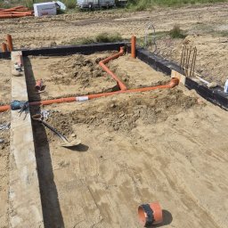 Murtechnik Paweł Żurek - Wykop pod konstrukcję budynku z ułożonymi pomarańczowymi rurami kanalizacyjnymi w piaszczystym gruncie, widoczne betonowe ławy i narzędzia.