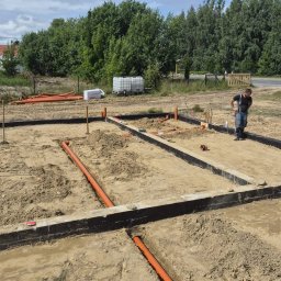Murtechnik Paweł Żurek - Przygotowany teren pod budowę z widocznymi, niskimi ścianami fundamentowymi i ułożonymi pomarańczowymi rurami drenażowymi w piasku. Pracownik nadzoruje postęp prac. W tle zieleń i błękitne niebo.