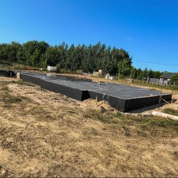 Murtechnik Paweł Żurek - Świeżo wylana płyta fundamentowa z czarną izolacją pionową, z wystającymi prętami zbrojeniowymi. Obok stoi betoniarka i kilku pracowników na tle lasu pod błękitnym niebem.