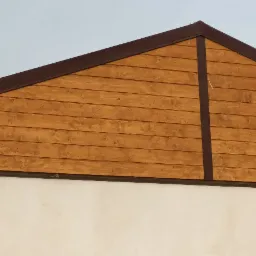 Szczyt domu z drewnianą elewacją w odcieniach brązu, wykończony ciemnobrązowymi listwami, nad białym murem.