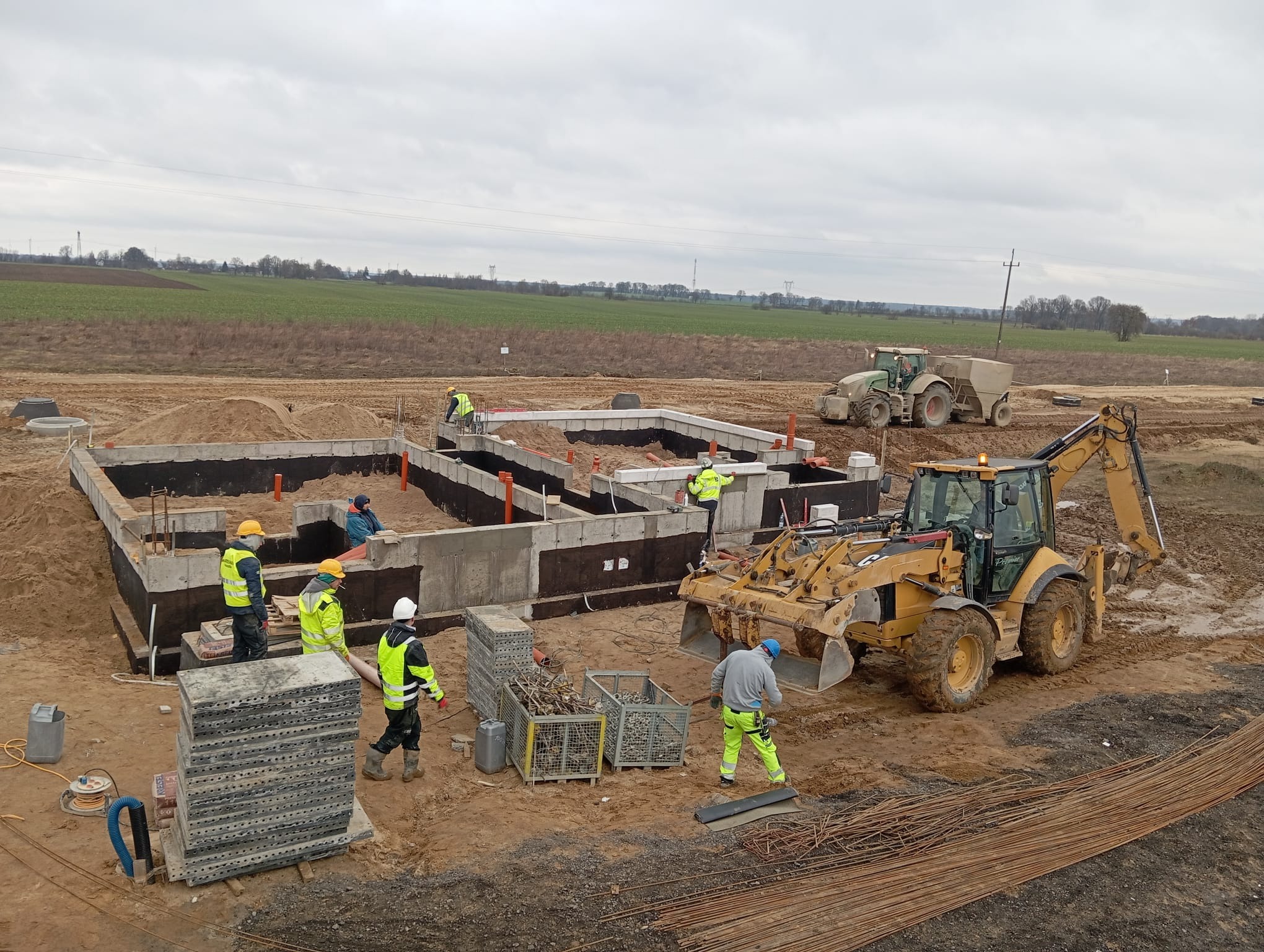 Budowa fundamentów: ekipa budowlana w żółtych kamizelkach pracuje przy betonowym fundamencie, widoczny traktor i koparka na placu budowy, ziemia wokół.