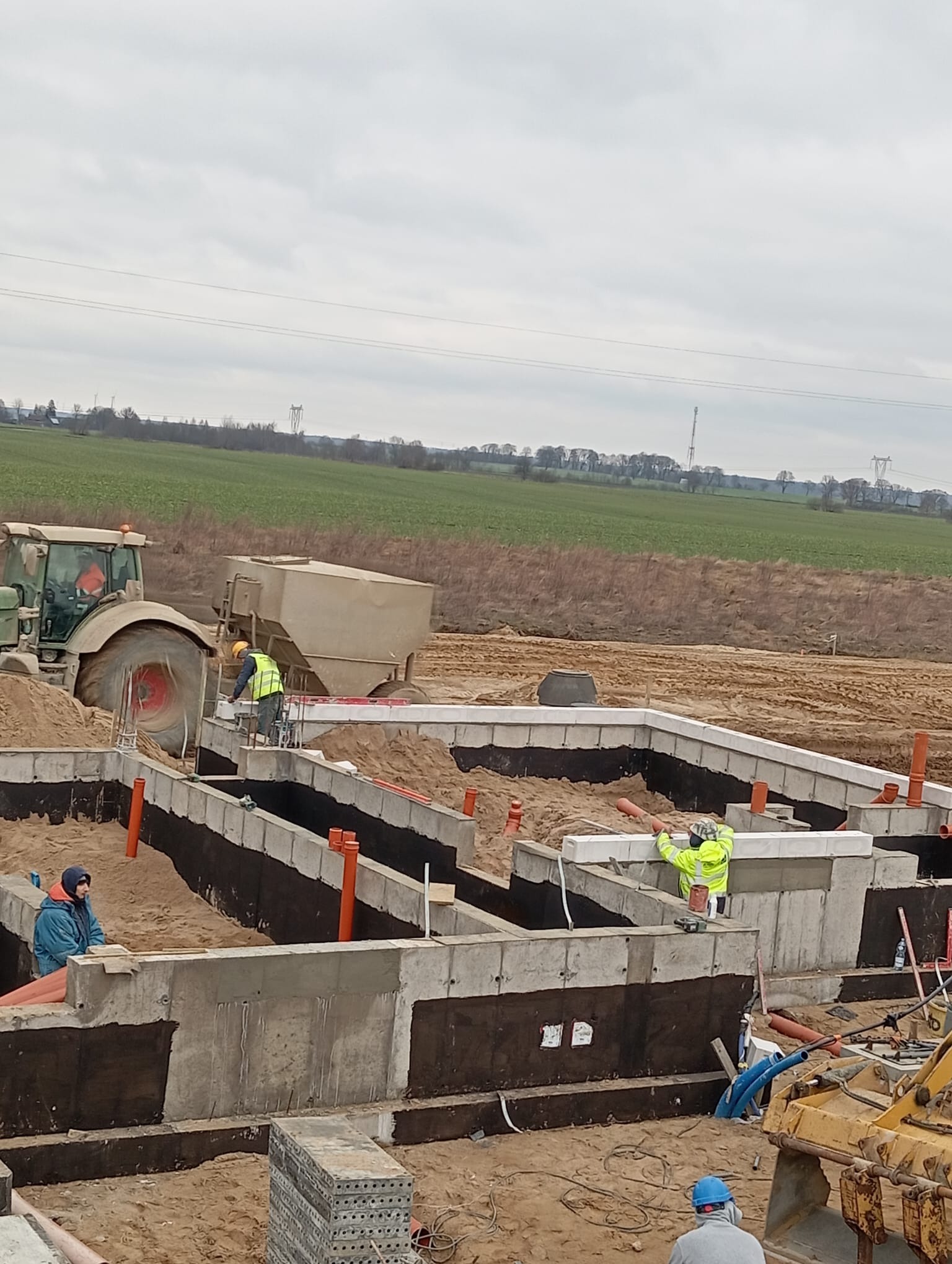 Budowa fundamentów domu jednorodzinnego: widok na wykopane i zabezpieczone ściany fundamentowe, pracownicy w odblaskowych kamizelkach, traktor z przyczepą na tle krajobrazu wiejskiego.
