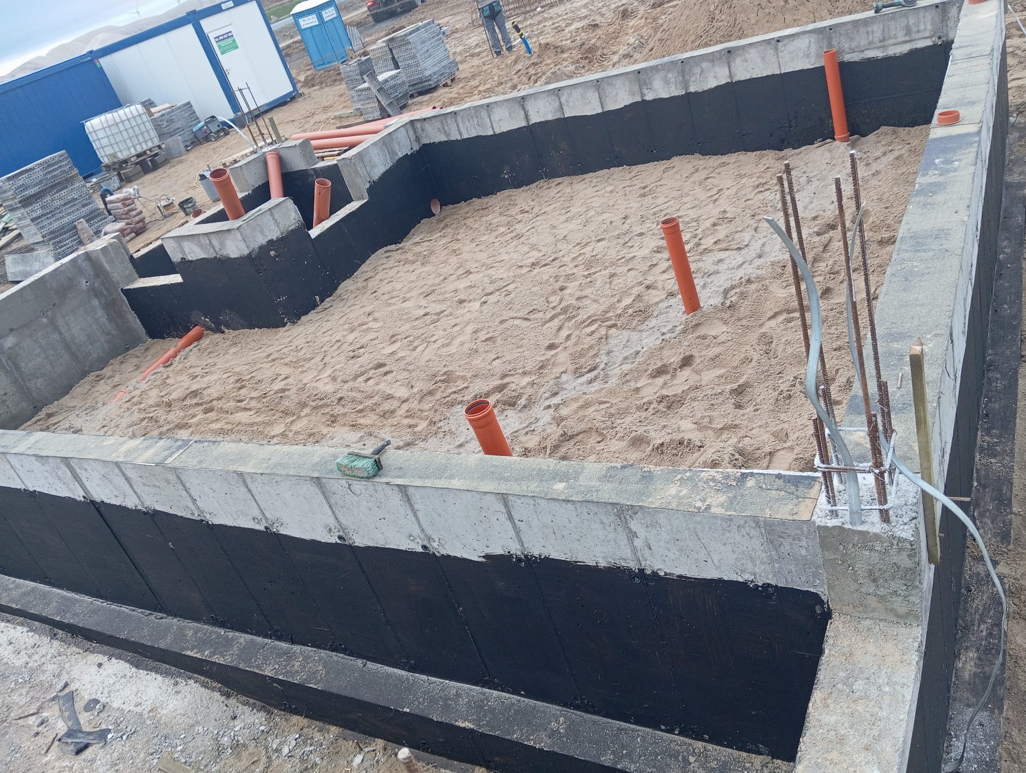 Wykopany fundament z izolacją bitumiczną, wypełniony piaskiem, z wystającymi rurami kanalizacyjnymi i prętami zbrojeniowymi, widoczne kontenery budowlane w tle.