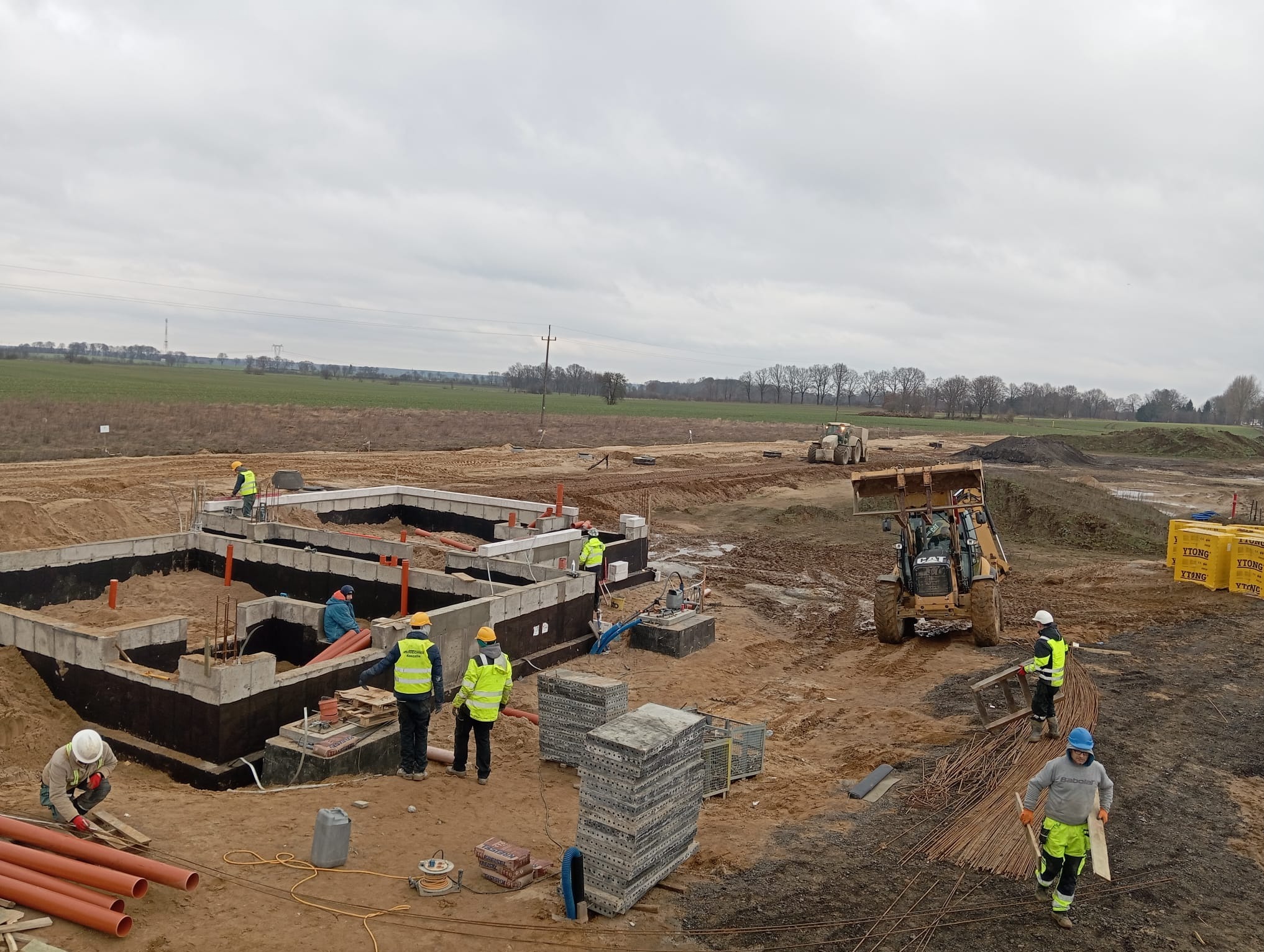 Szeroki kadr z budowy fundamentów domu: pracownicy w odblaskowych kamizelkach, wykopane fundamenty z izolacją, rury kanalizacyjne, sterta zbrojenia i koparko-ładowarka CAT w tle, pochmurne niebo.
