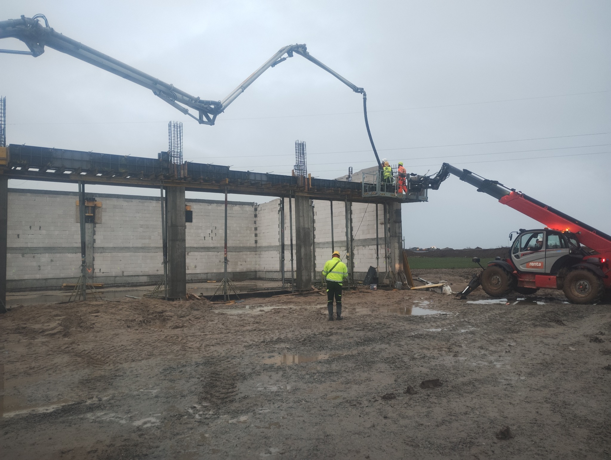 Wylewanie betonu na strop budynku z betonu komórkowego. Na górze pracownicy, widoczny wysięgnik pompy do betonu i teleskopowa ładowarka. Teren błotnisty.