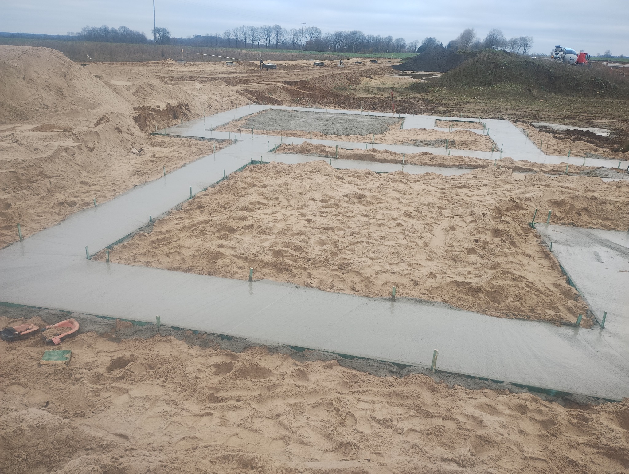 Wylane fundamenty pod budowę domu jednorodzinnego, widoczne szalunki i świeży beton, teren piaszczysty, w tle gruszka do betonu.