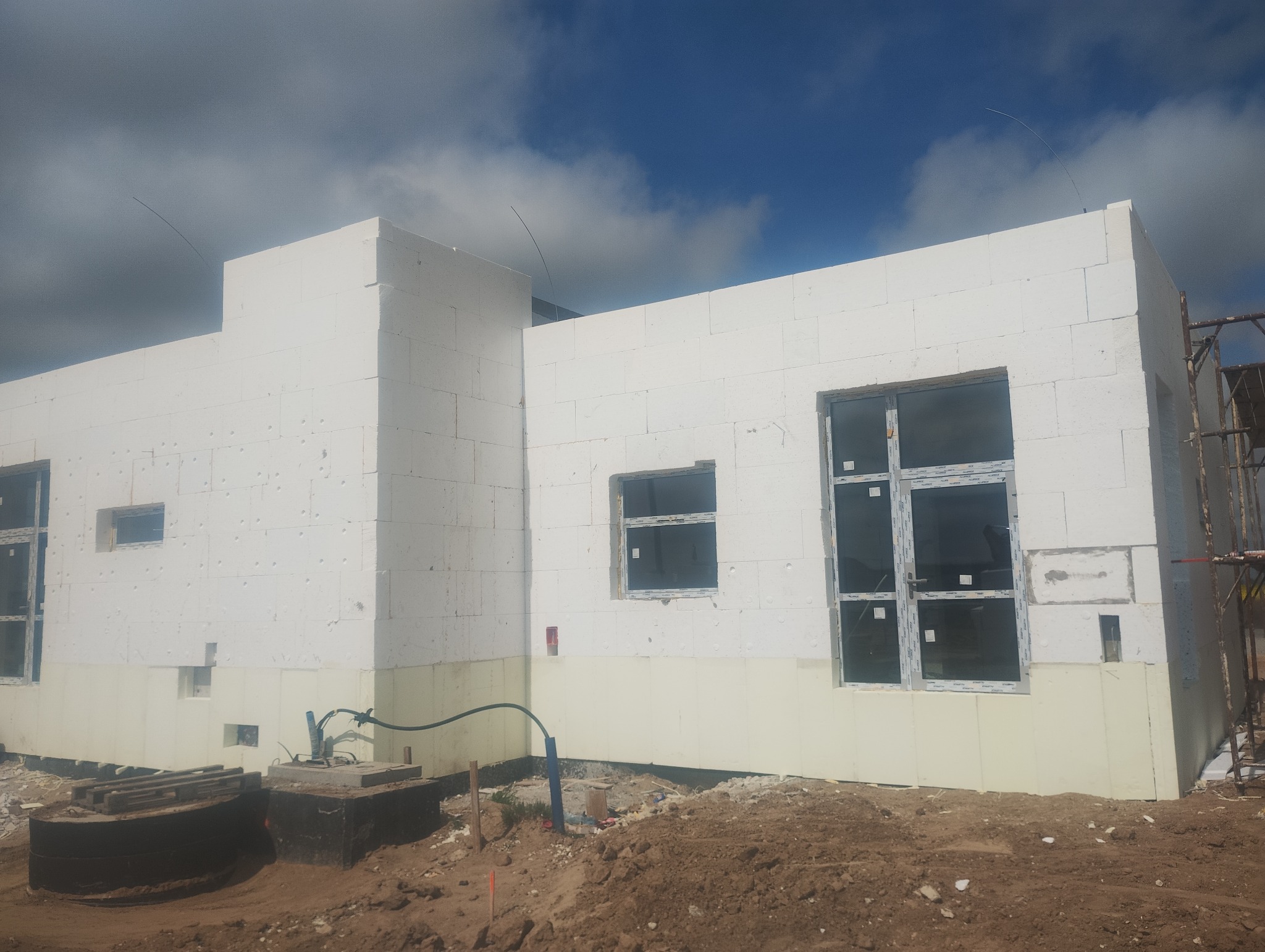 Budowa domu z białych bloczków, widoczne okna z folią ochronną, ocieplenie fundamentów. Ujęcie w słoneczny dzień, na pierwszym planie ziemia.