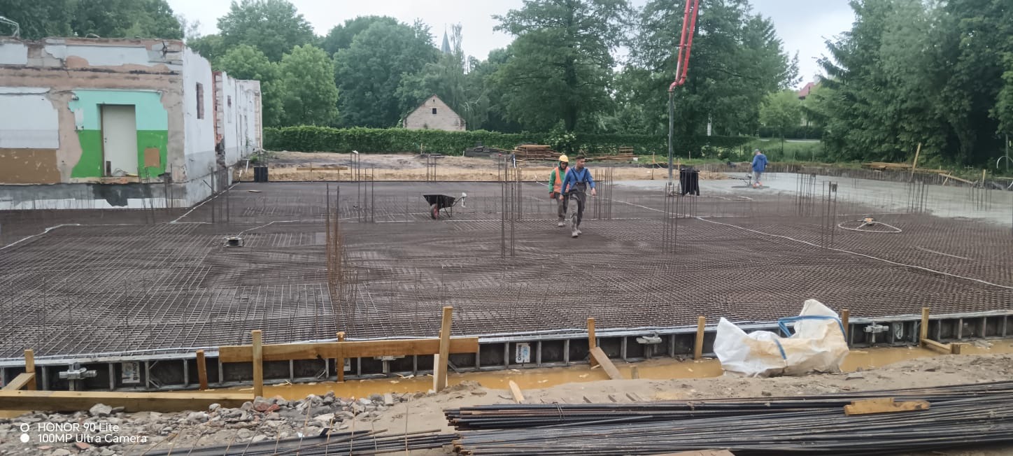 Zbrojenie płyty fundamentowej pod budynek z częściowo rozebraną ścianą. Na zbrojeniu pracownicy budowlani i pompa do betonu w tle. Widoczne szalunki i pręty zbrojeniowe.