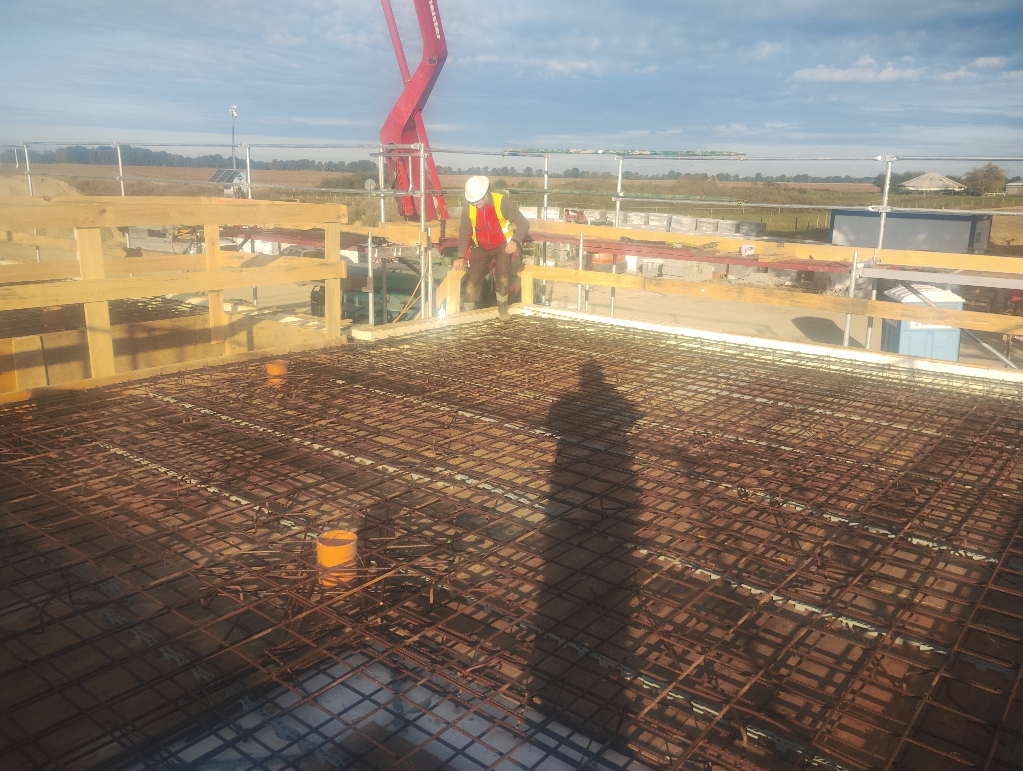 Zbrojenie płyty fundamentowej z widocznym pracownikiem w kamizelce i elementami szalunku, przygotowane do zalewania betonem. Widok z góry, w tle krajobraz.