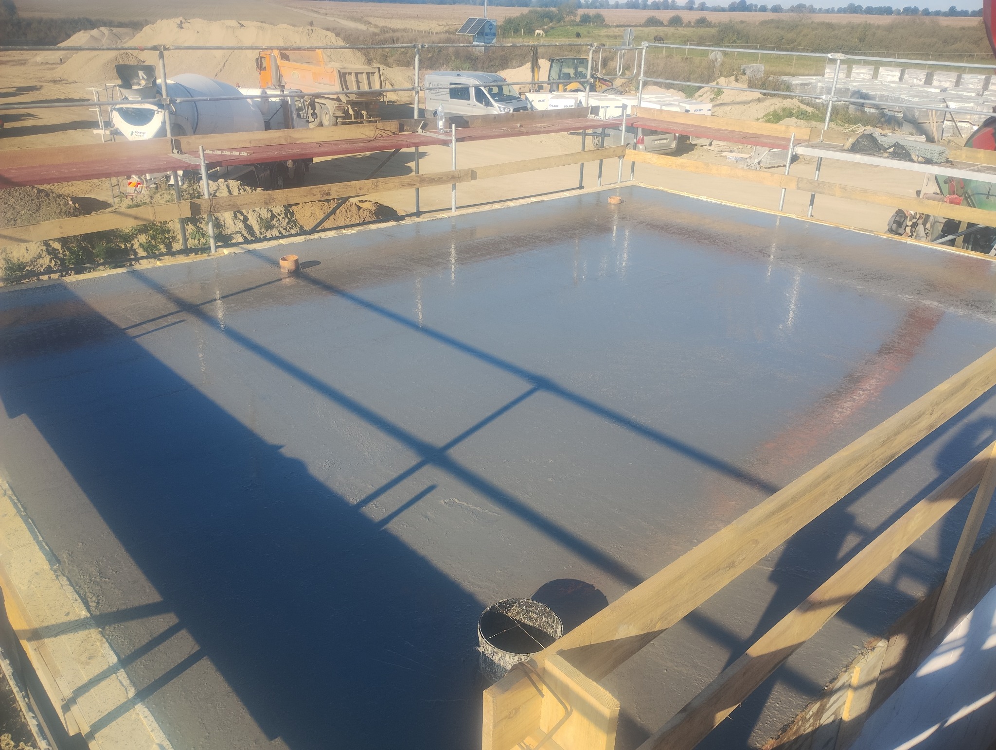 Świeżo wylana płyta fundamentowa z widocznymi elementami szalunku i instalacji. W tle maszyny budowlane i hałda piasku. Słoneczny dzień, plac budowy.