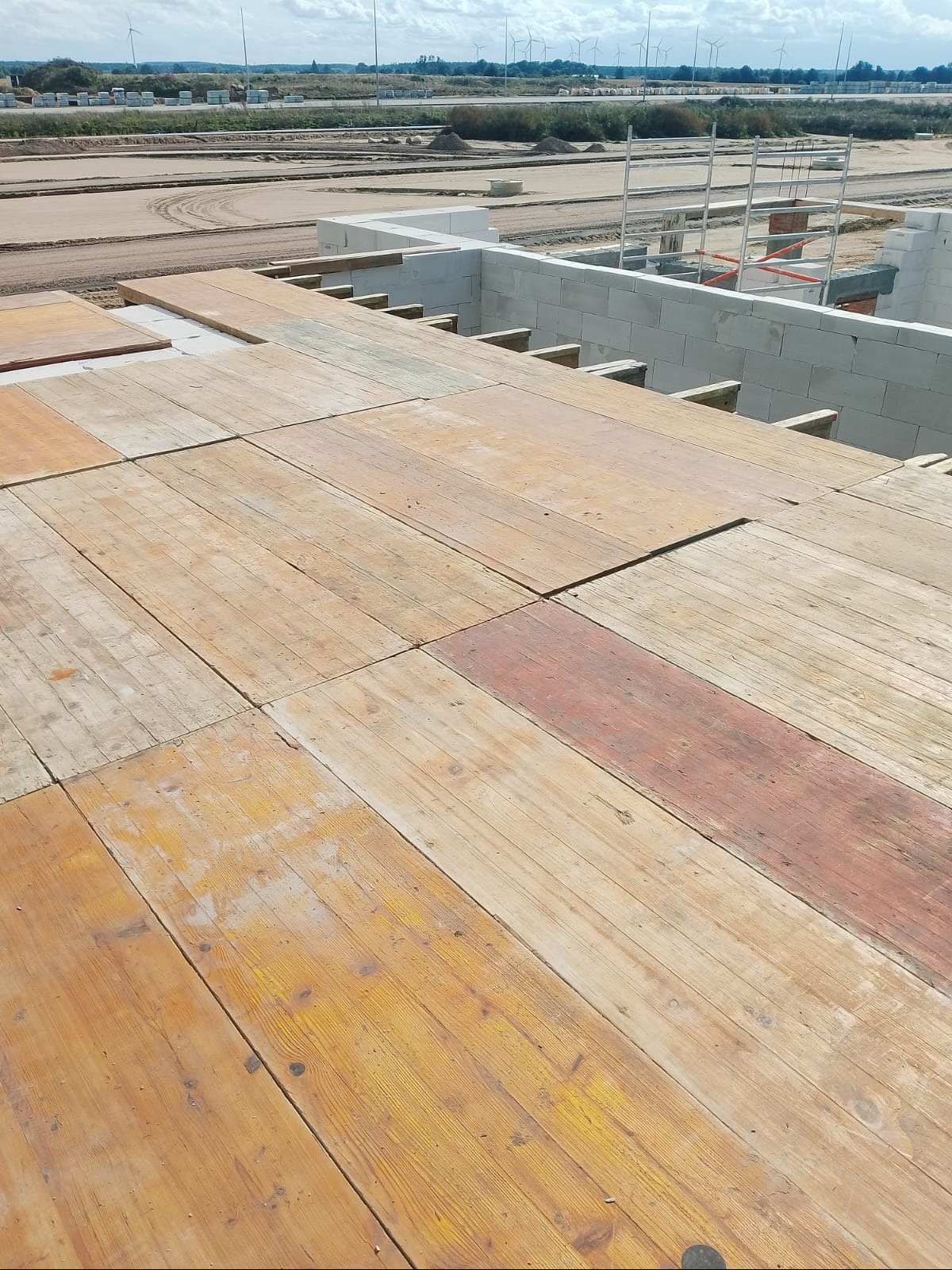 Wysoki kąt na drewniany szalunek stropowy na budowie z bloczków, z widocznymi turbinami w oddali. Widok na plac budowy z elementami konstrukcyjnymi.