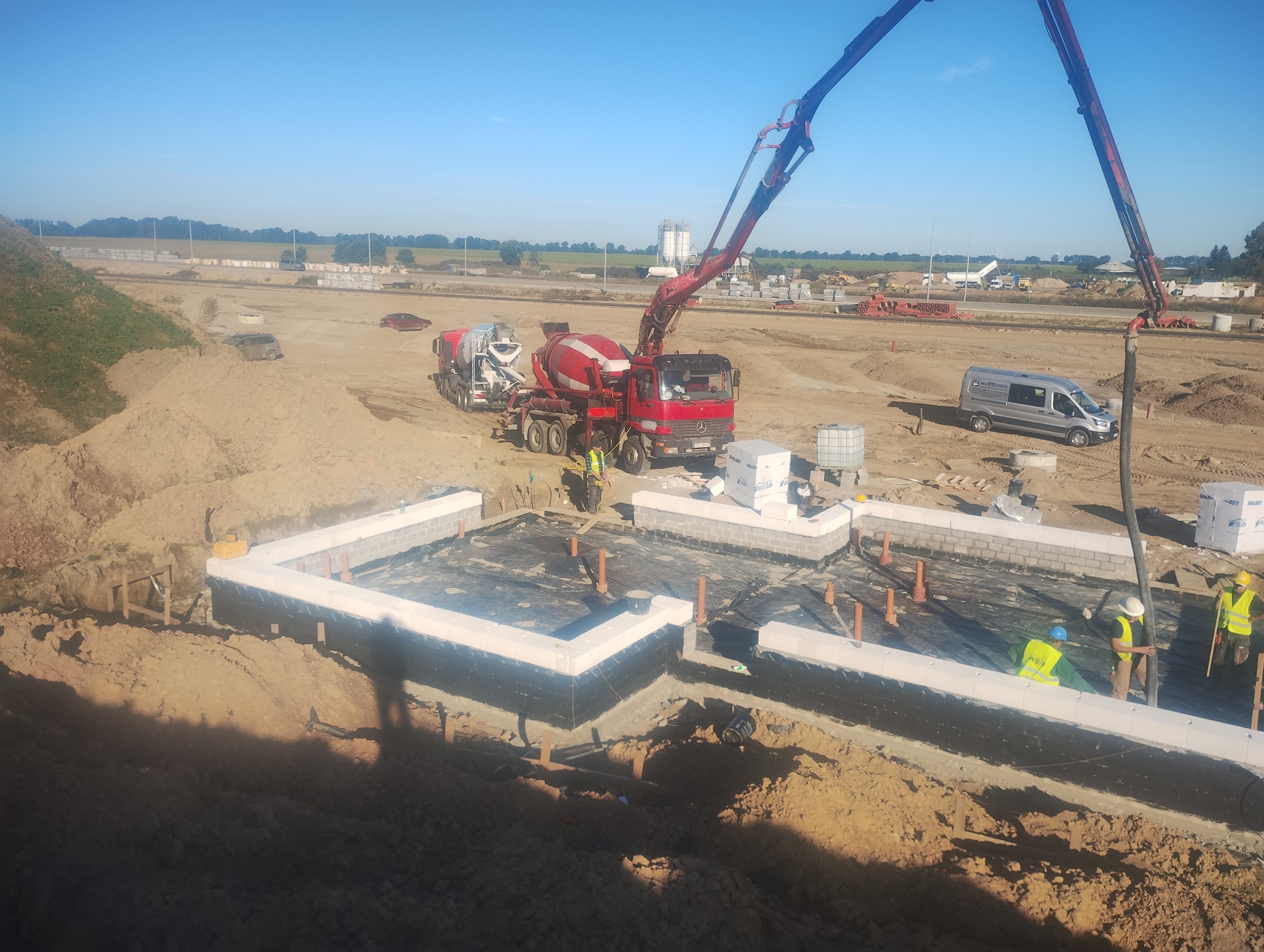 Wykop pod fundamenty z hydroizolacją, w tle betoniarka z pompą podająca beton. Widoczni pracownicy w odblaskowych kamizelkach. Słoneczny dzień na placu budowy.