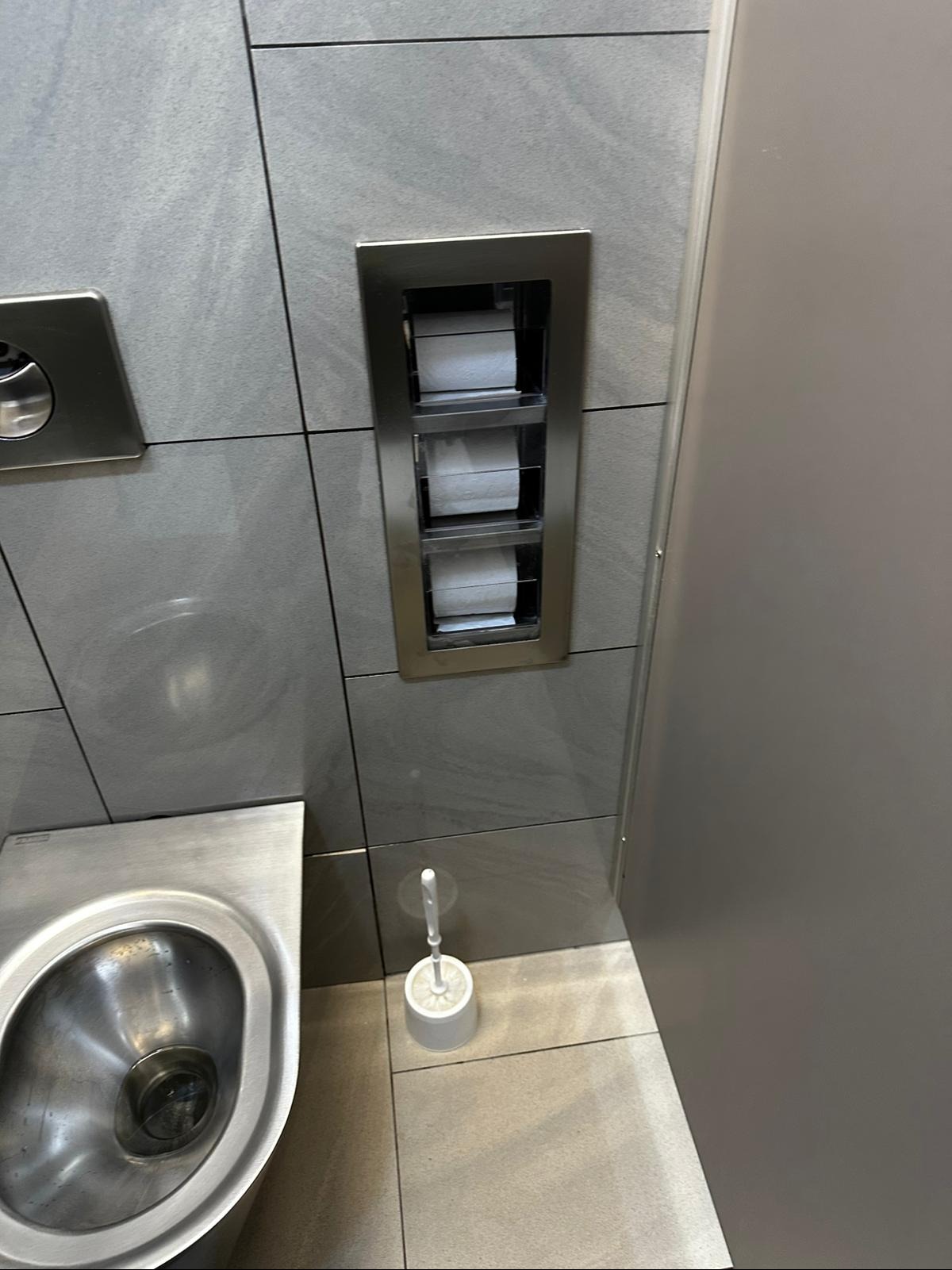 Nowoczesna łazienka z metalową miską WC, wbudowanym pojemnikiem na papier toaletowy i szczotką. Szare płytki na ścianach i podłodze. Minimalistyczny design.