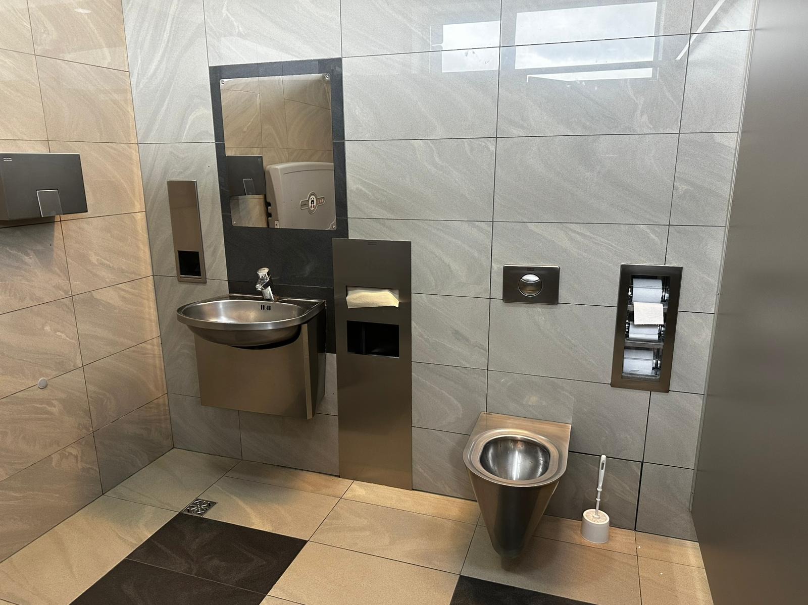 Nowoczesna łazienka publiczna z umywalką, toaletą ze stali nierdzewnej i dozownikiem papieru. Płytki imitujące kamień w odcieniach szarości. Minimalistyczny design.