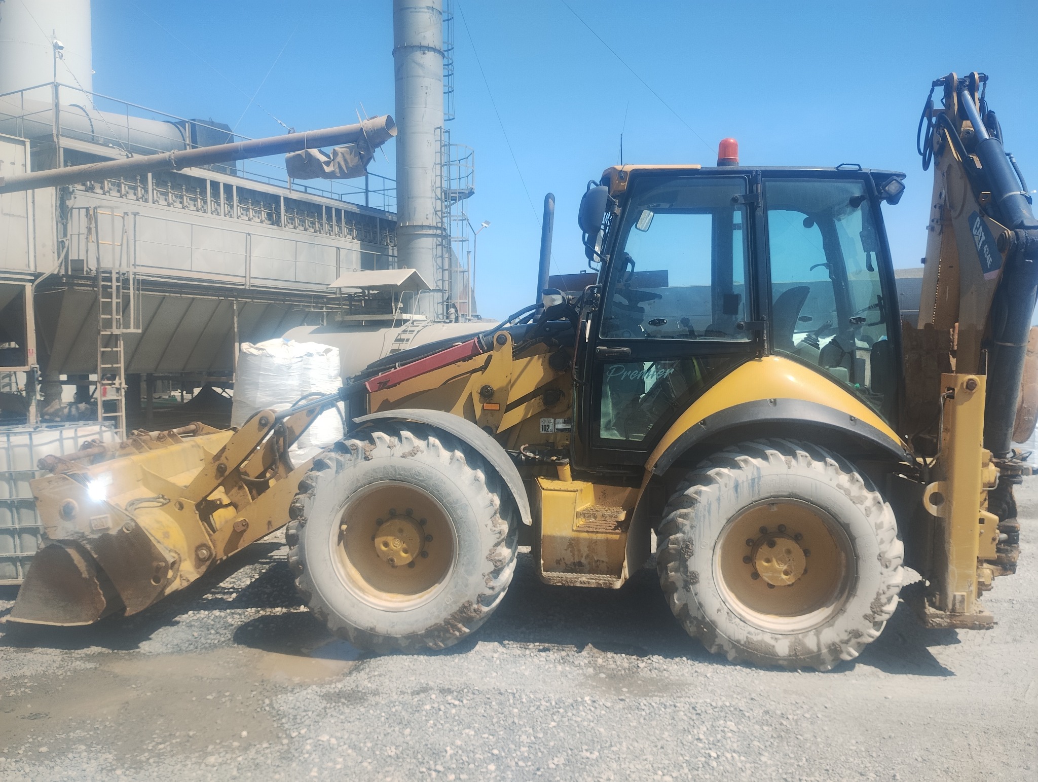 Żółta koparko-ładowarka na tle zakładu przemysłowego w słoneczny dzień. Widoczne duże opony i kabina z napisem 'Premier'.