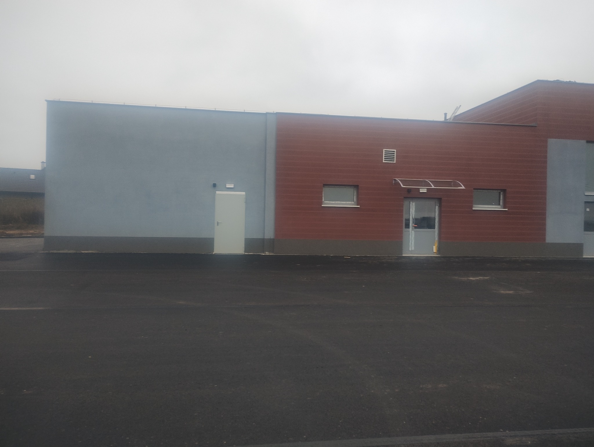 Nowoczesny budynek z dwukolorową elewacją: szara i ceglana. Widoczne okna, drzwi wejściowe z daszkiem i fragment dachu. Asfaltowy plac przed budynkiem.