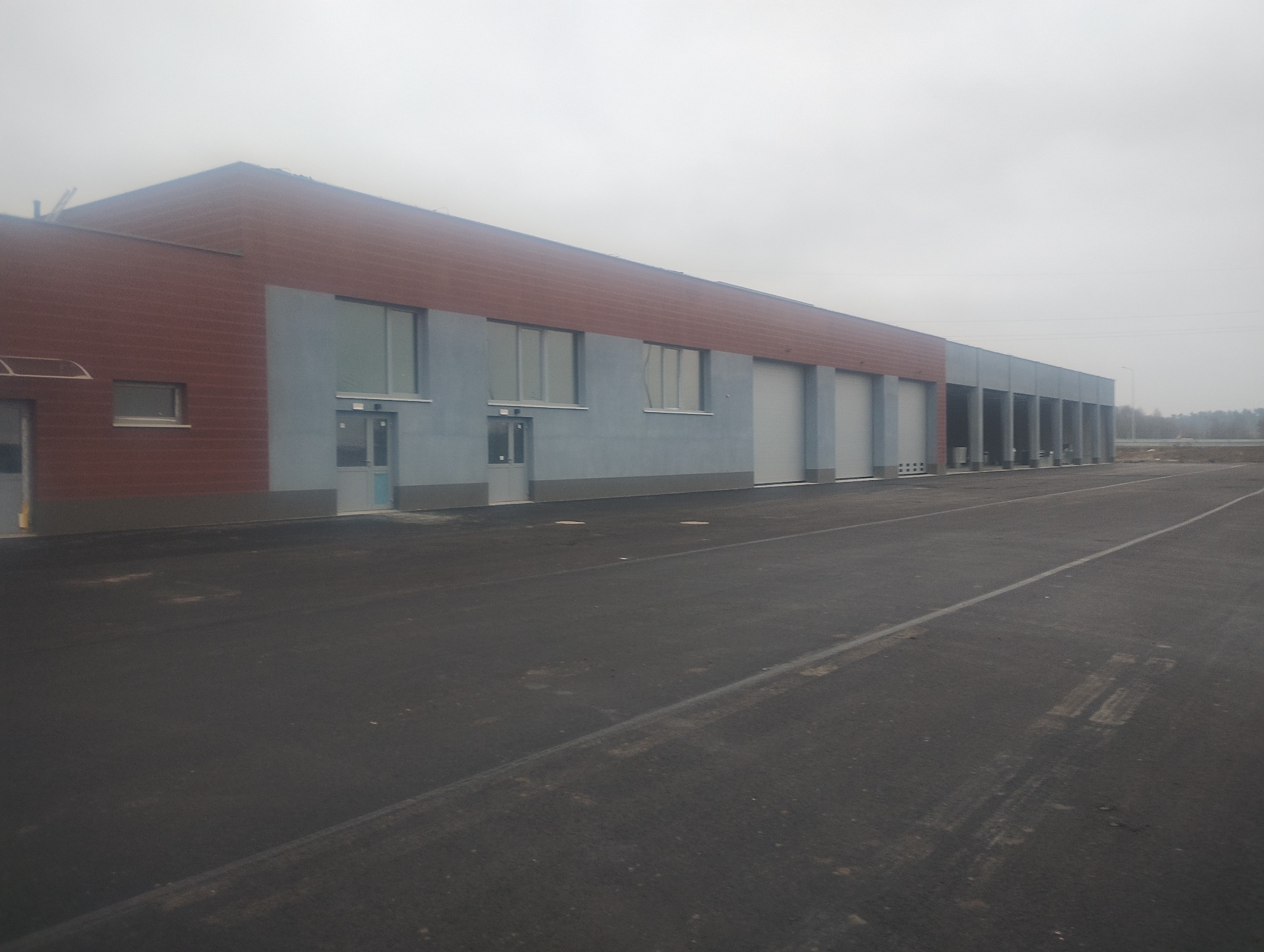 Nowoczesny budynek przemysłowy z szarą elewacją i segmentowymi bramami garażowymi, widoczny z perspektywy placu manewrowego w pochmurny dzień.