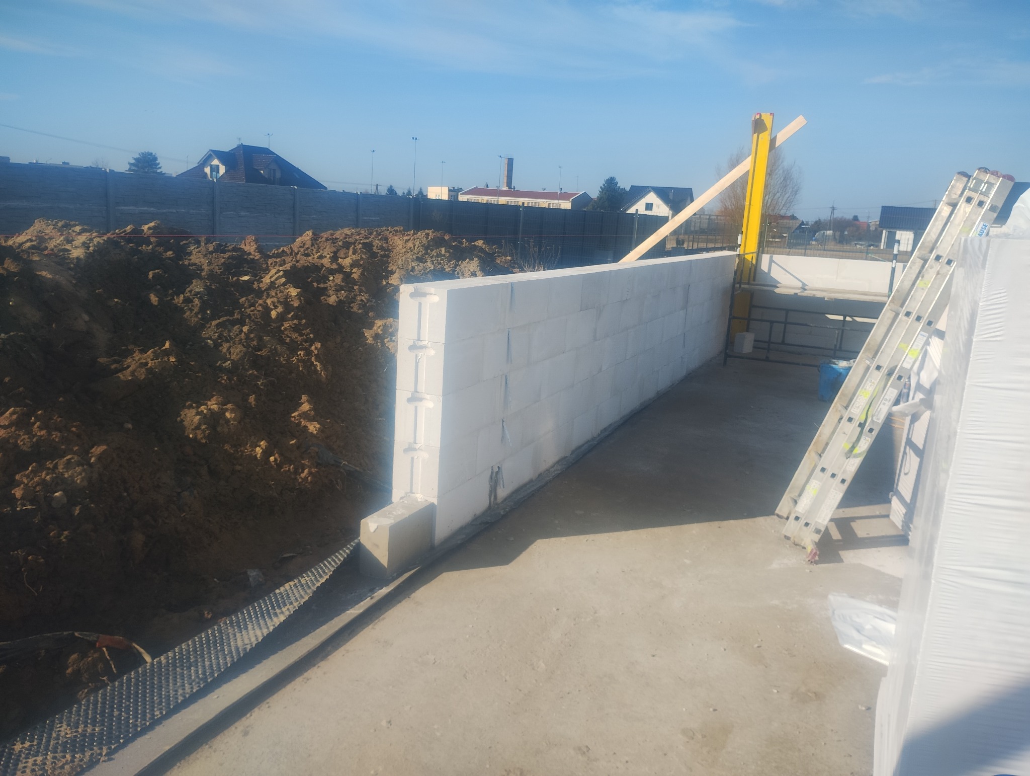 Wznoszenie ściany z białych bloczków betonowych na fundamencie. Obok leży pryzma ziemi i stoi drabina. Widoczne elementy szalunku. Słoneczny dzień.