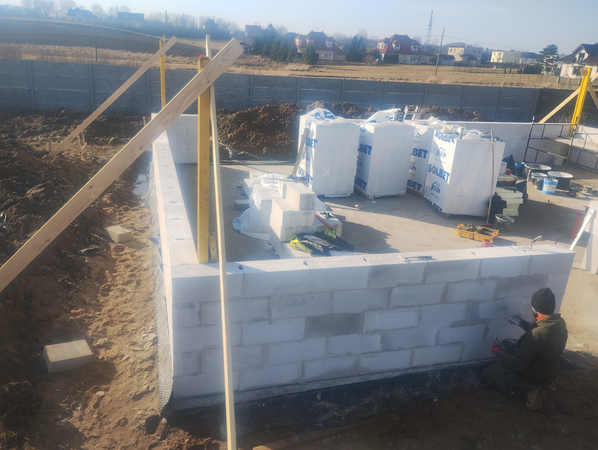 Wznoszenie ścian fundamentowych z bloczków Solbet. Widoczne narzędzia, poziomica i zapas materiałów budowlanych. Prace murarskie w toku, zarys przyszłego budynku.