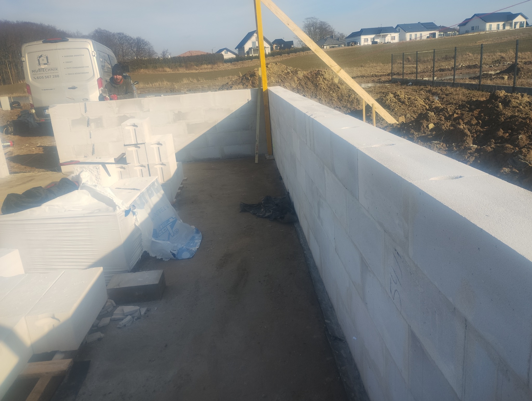 Wznoszenie ścian fundamentowych z bloczków betonu komórkowego. Widoczny fragment ściany, poziomica, palety bloczków i samochód dostawczy na tle krajobrazu.
