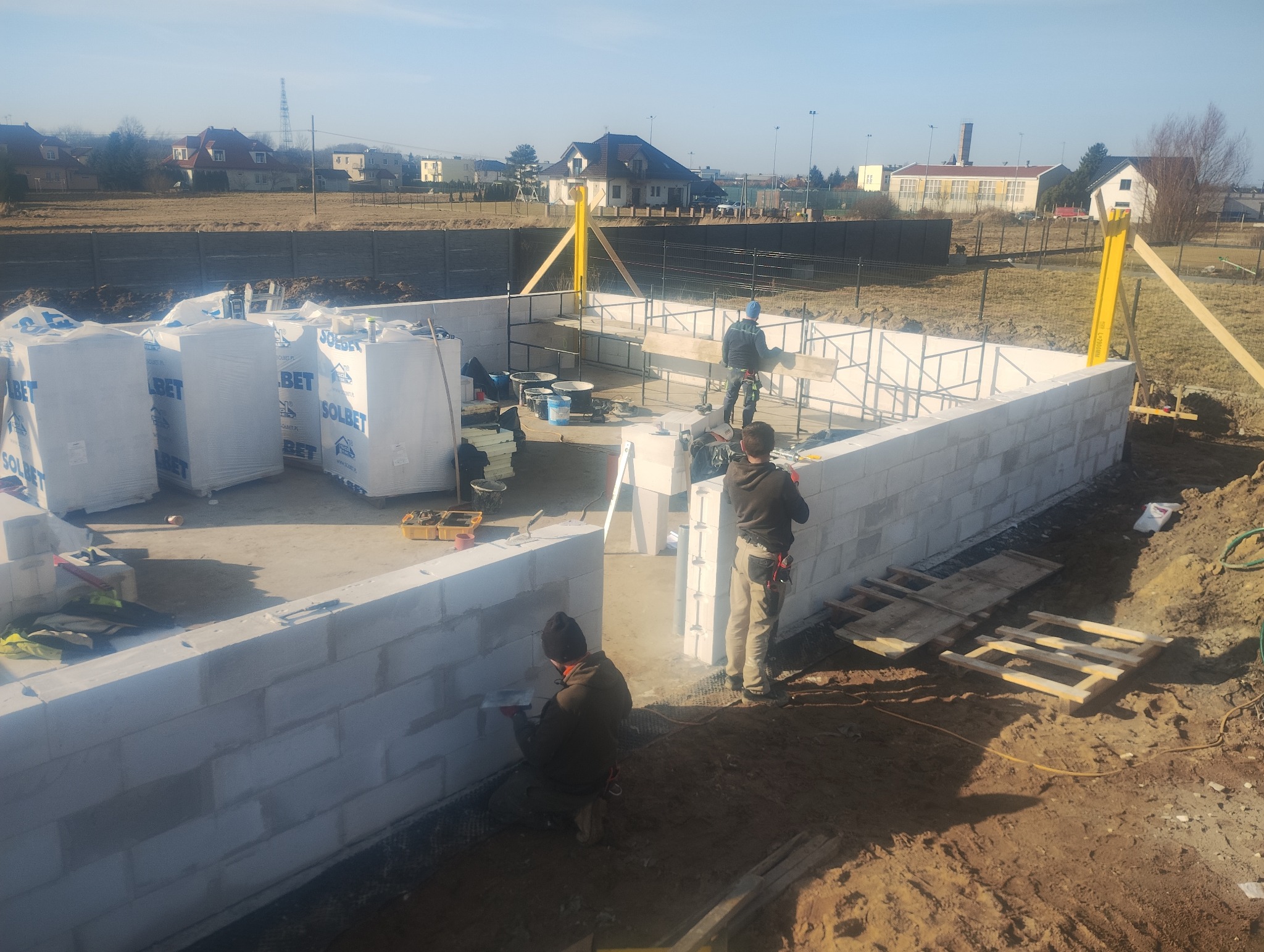 Wznoszenie ścian fundamentowych z bloczków Solbet. Widoczni pracownicy budowlani, narzędzia i materiały budowlane. W tle zabudowa jednorodzinna.