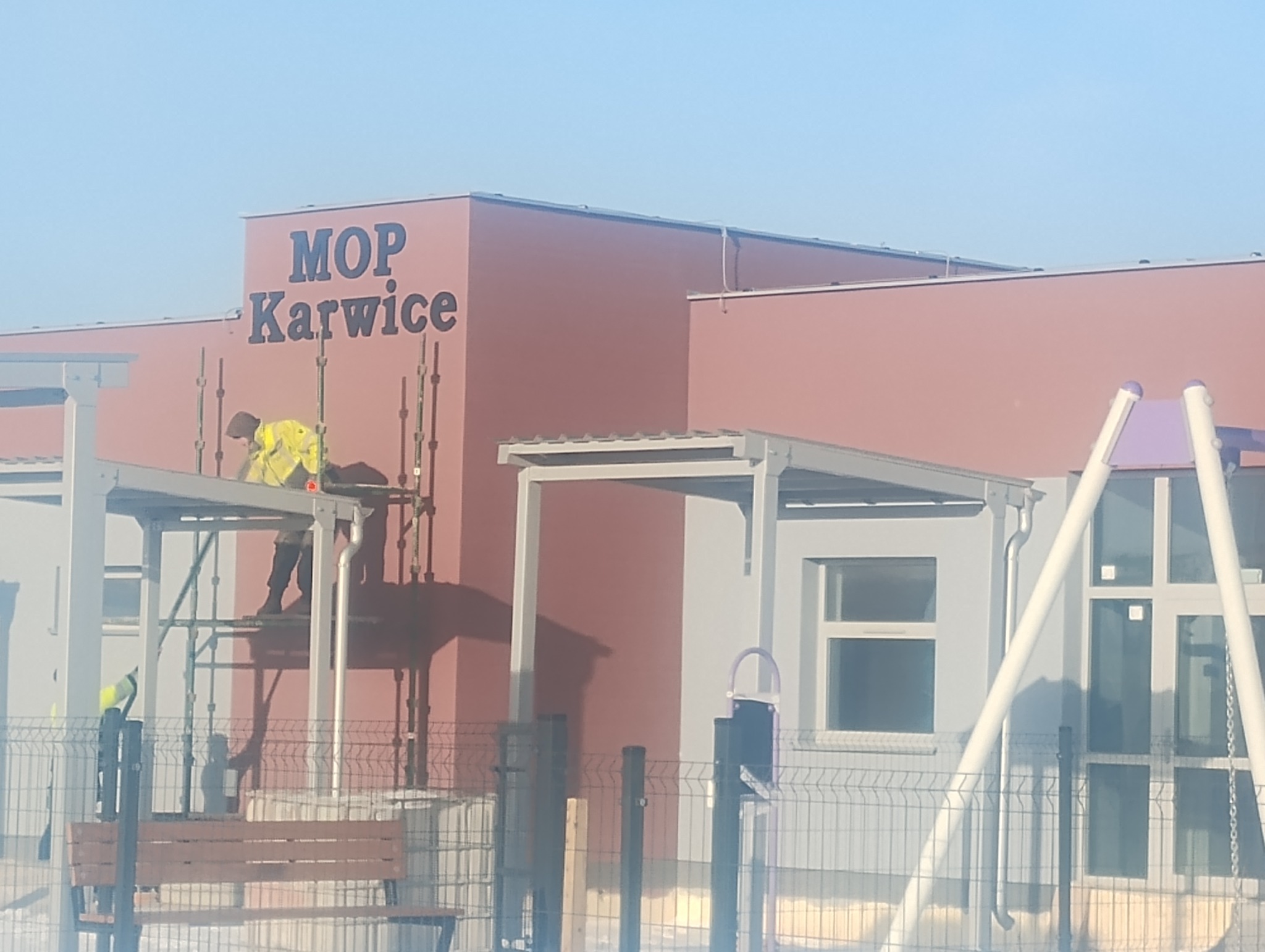 Pracownik w żółtej kamizelce na rusztowaniu przy budynku MOP Karwice. Montaż elewacji lub prace wykończeniowe. Widoczne fragmenty placu zabaw.