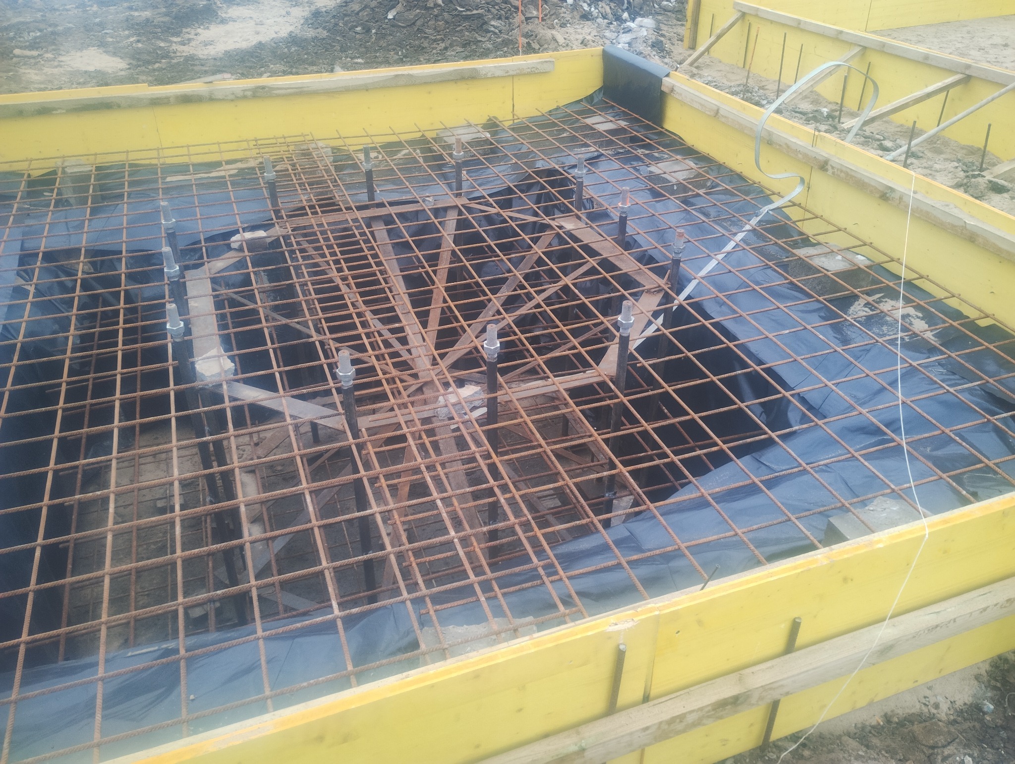 Zbrojenie fundamentu z żółtym szalunkiem i czarną folią hydroizolacyjną. Widoczne wystające pręty z gwintem. Ujęcie z góry, ukazujące siatkę zbrojeniową.