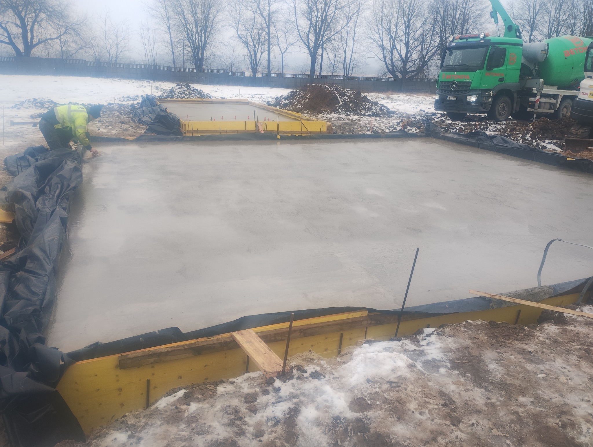 Wylana płyta fundamentowa z żółtym szalunkiem i czarną folią, na zewnątrz pracownik wyrównuje beton. W tle zielona betoniarka i zasypana śniegiem ziemia.