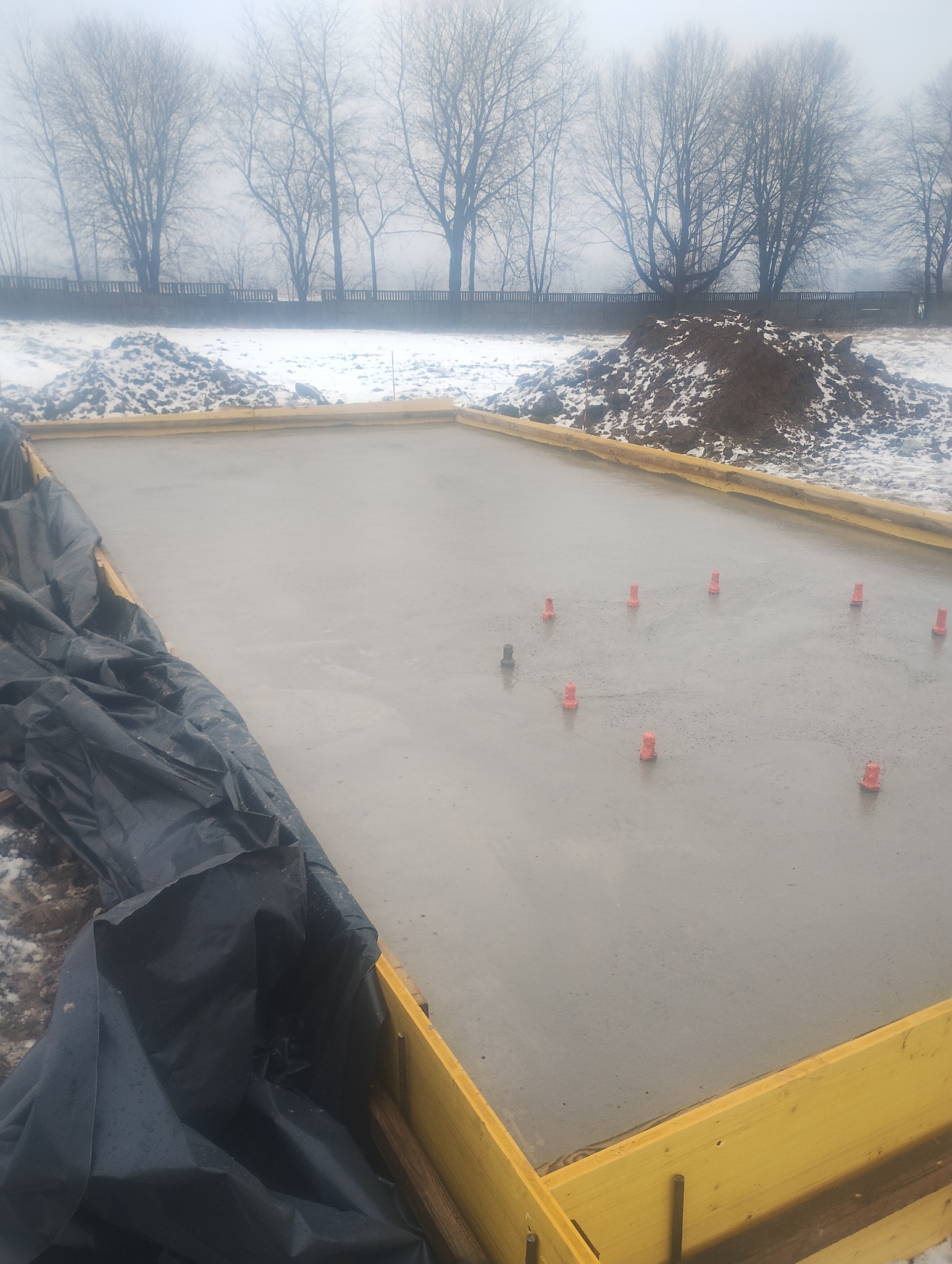 Wylana, świeża płyta fundamentowa w drewnianym szalunku, z widocznymi elementami poziomującymi. W tle zasypana ziemia i drzewa w zimowej scenerii.