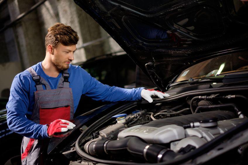 Mechanik w kombinezonie roboczym sprawdza silnik samochodu z otwartą maską, używając rękawic ochronnych.