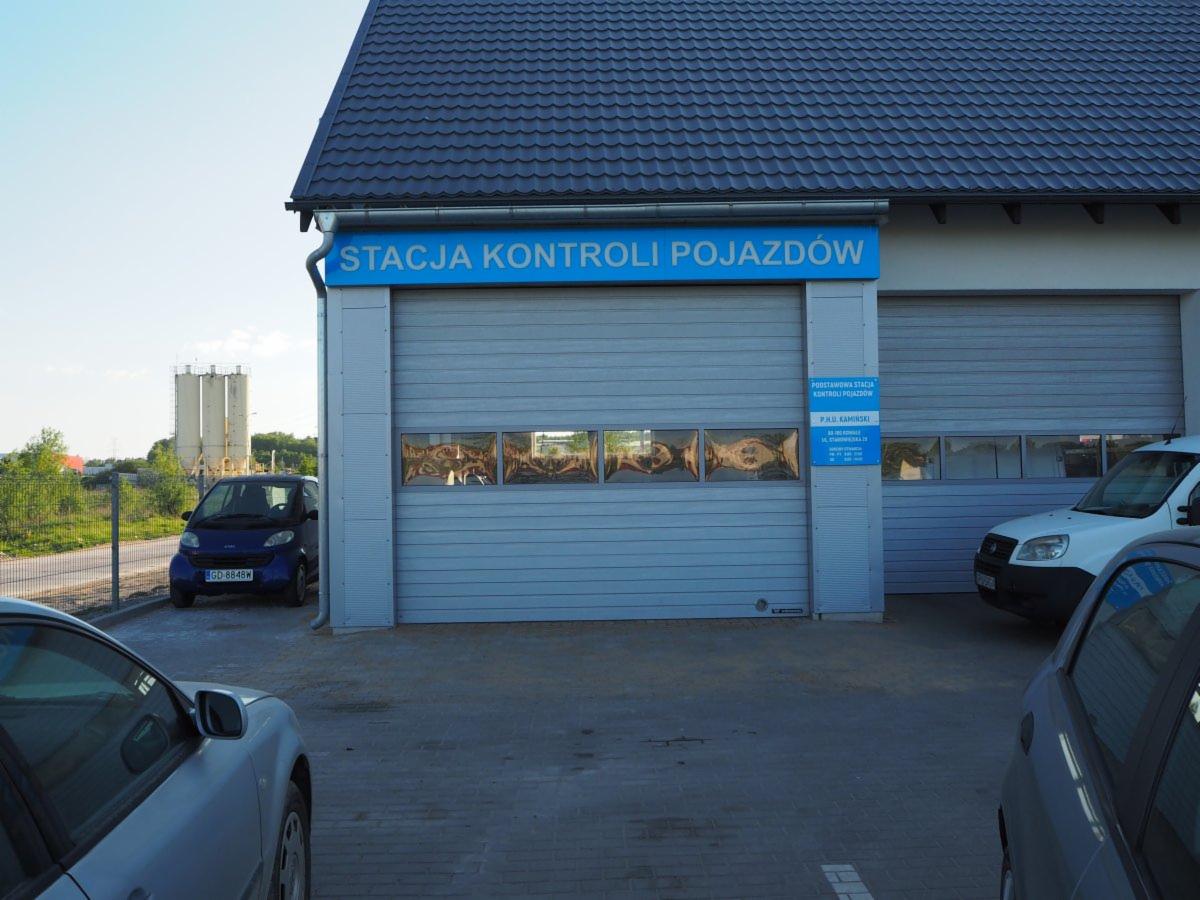 Zewnętrzny widok stacji kontroli pojazdów z niebieskim szyldem, zaparkowanym niebieskim Smartem i częściowo widocznymi samochodami osobowymi na placu przed wjazdem.