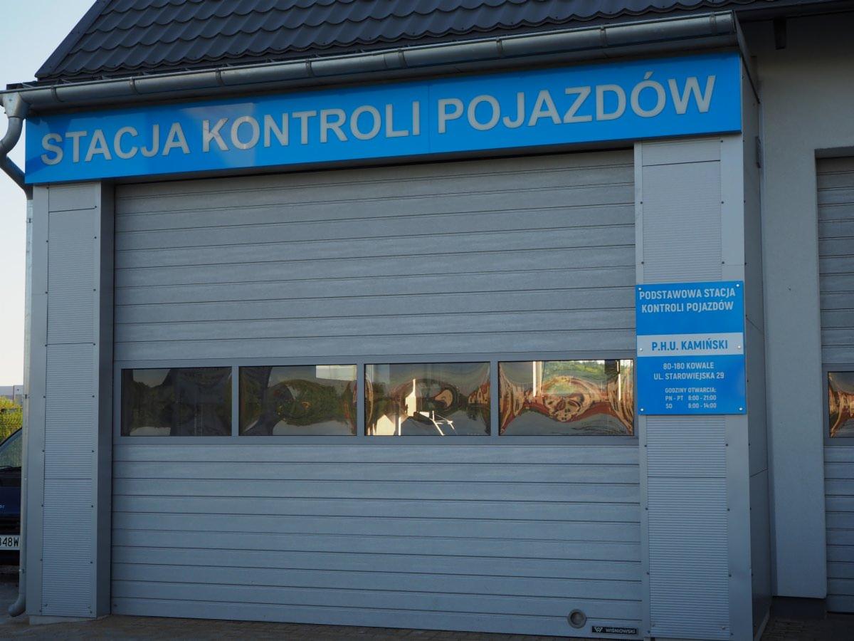 Budynek stacji kontroli pojazdów z szarymi rolowanymi drzwiami garażowymi i niebieskim szyldem z napisem 'STACJA KONTROLI POJAZDÓW', widoczny również mniejszy szyld informacyjny z danymi firmy.