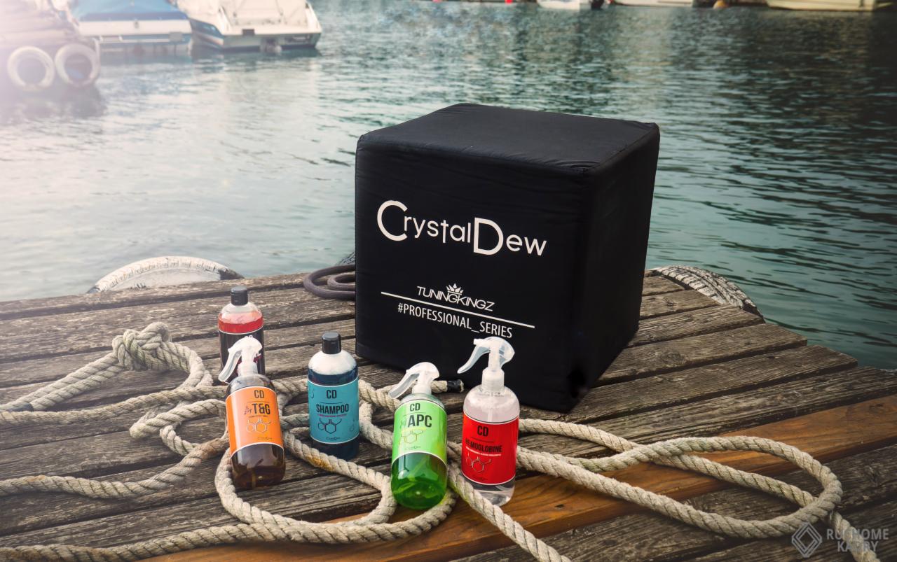 Zestaw kosmetyków samochodowych Crystal Dew na drewnianym pomoście z liną, na tle wody i zacumowanych łodzi.
