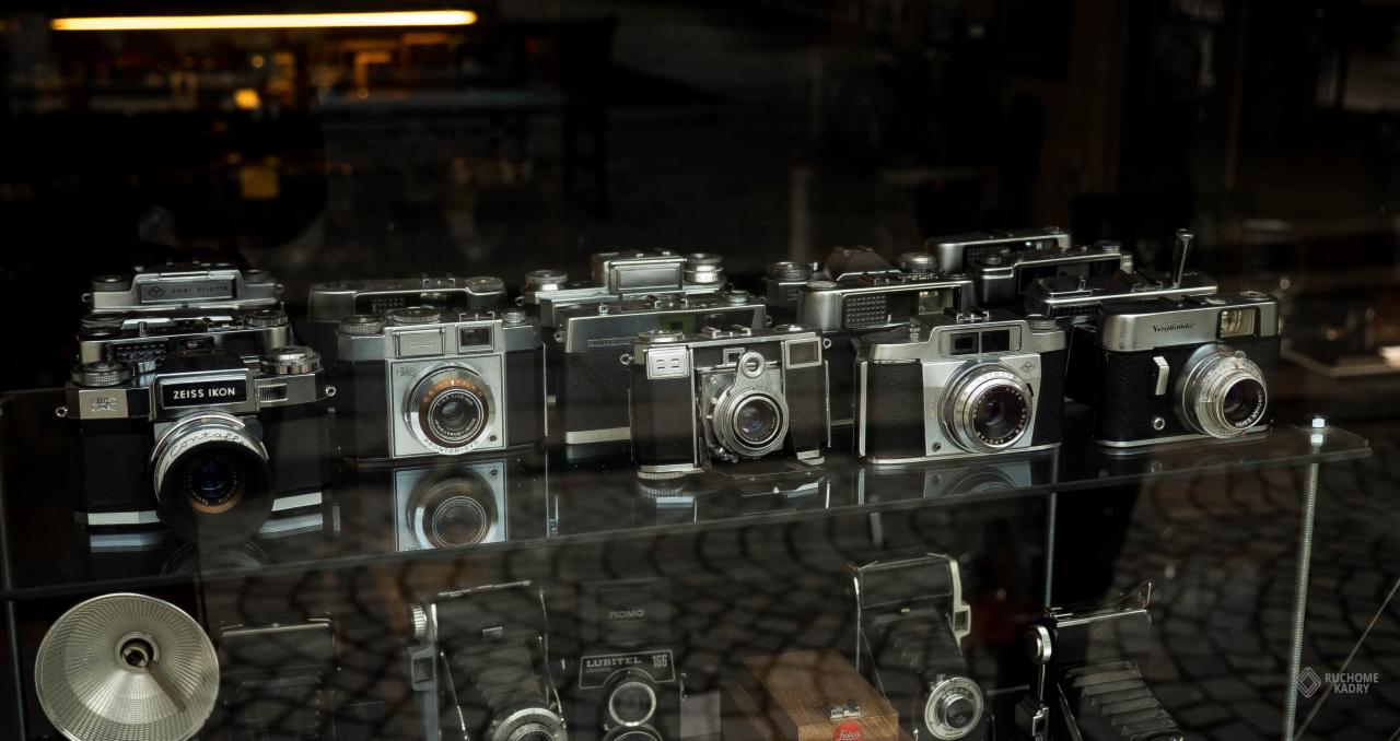 Kolekcja zabytkowych aparatów fotograficznych różnych marek, eksponowanych na szklanej półce w witrynie sklepowej, z widocznym odbiciem wnętrza sklepu w szybie.