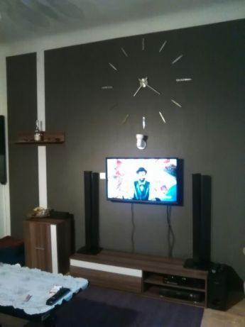 Ciemnoszara ściana z naklejanym zegarem, telewizorem, głośnikami i półką z butelką alkoholu, po lewej stronie pionowy biały pas.