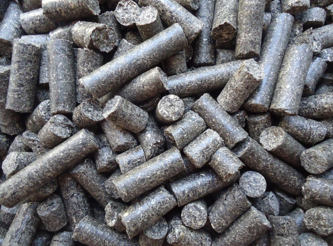 Stos ciemnoszarych cylindrycznych brykietów z biomasy, widoczne nieregularne pęknięcia i tekstura włókien, oświetlenie równomierne.