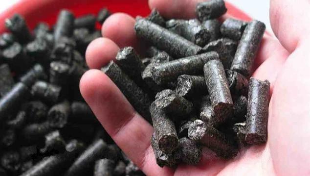 Garść ciemnych, cylindrycznych brykietów opału, prawdopodobnie z biomasy, trzymana w dłoni na tle czerwonego pojemnika z większą ilością tego samego materiału.