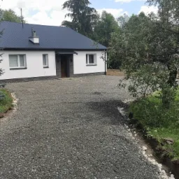 Szary żwirowy podjazd prowadzący do białego domu z ciemnym dachem, widoczne obrzeża i przygotowany teren pod trawnik.