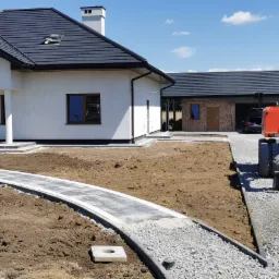 Ułożona ścieżka z kostki brukowej w kształcie łuku prowadząca do nowego domu z białymi ścianami i ciemnym dachem, pracownik wygładza ziemię, w tle pomarańczowa koparka i budynek gospodarczy z cegły.